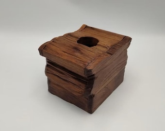 May include: A handcrafted wooden tissue box with a square shape and a dark brown finish. The box has a rough, textured surface and a circular opening on top for dispensing tissues. The design gives a rustic, natural aesthetic.