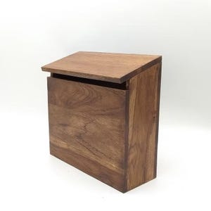 Könnte beinhalten: Ein dunkelbrauner Holzbriefkasten mit schrägem Deckel und rechteckigem Korpus. Die Holzmaserung ist sichtbar. Der Deckel ist leicht geöffnet und gibt den Blick ins Innere frei.