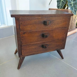 May include: A small, vintage wooden nightstand with three drawers. The nightstand is made of dark brown wood and features a rectangular top, three drawers with small, curved handles, and angled legs. The wood grain is visible.