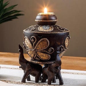 Puede incluir: Un portavelas decorativo con un acabado marrón oscuro y detalles tejidos intrincados. El portavelas presenta un diseño de flor tallada y está sostenido por dos figuras de elefantes.