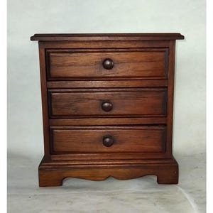 May include: A small wooden chest of drawers with three drawers. The drawers are made of dark wood and have simple metal knobs. The chest is sitting on a white surface.