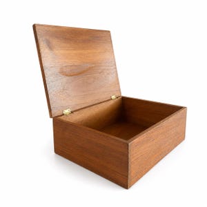 May include: A rectangular wooden box with a natural brown wood grain finish and an open hinged lid. The box features two small gold-toned metal hinges connecting the lid to the base, showcasing a simple, clean construction with visible wood texture.
