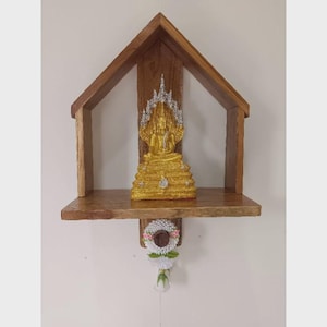 May include: A wooden shelf in the shape of a house, with a golden Buddha statue and a floral garland. The Buddha is seated and has a silver and gold halo. The shelf is made of dark wood and is mounted on a white wall.