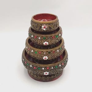 May include: A stack of five decorative wooden bowls. The bowls are dark red inside and have ornate, bronze-colored metalwork around the rims. The metalwork is decorated with small, colorful gems and floral accents.