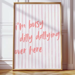 May include: A framed art print with a light wood frame. The artwork features a pink and white striped background with the text "I'm busy dilly dallying over here" in pink cursive lettering. The print is displayed against a white wall and wooden floor.