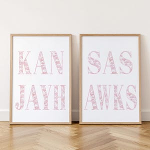 May include: Two vertical rectangular wall art prints in light wood frames. The left print displays the text KAN JAYH in large serif letters with a pink floral pattern. The right print displays the text SAS AWKS in the same pink floral style.
