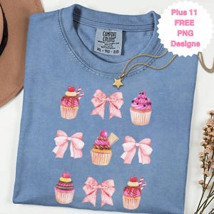May include: A light blue t-shirt with a crew neck, featuring a watercolor design of cupcakes and pink bows. The shirt has a Comfort Colors tag and a gold star necklace. The text "Plus 11 FREE PNG Designs" is also visible.
