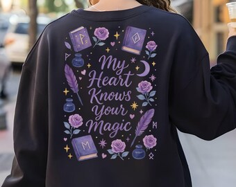 May include: A navy blue sweatshirt with a graphic design on the back. The design includes the text "My Heart Knows Your Magic" surrounded by purple roses, books, feathers, and stars.