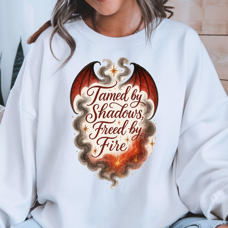 May include: White sweatshirt featuring a dragon wing design in red and black, with the text "Tamed by Shadows, Freed by Fire" in elegant script. The design includes smoke and star accents, creating a fantasy theme.