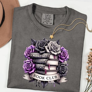 May include: A grey Comfort Colors t-shirt with a graphic of a stack of books and purple and grey roses. The books are labelled "Morally Grey" and the banner reads "Book Club". A gold star necklace is draped on the shirt.