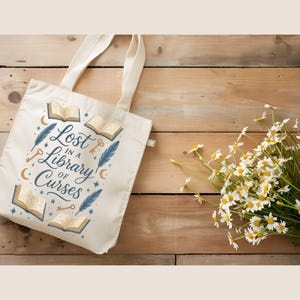 May include: Cream-colored tote bag with the text "Lost in a Library of Curses" and illustrations of books, keys, feathers, and stars. The bag is on a wooden surface next to a bouquet of white daisies.