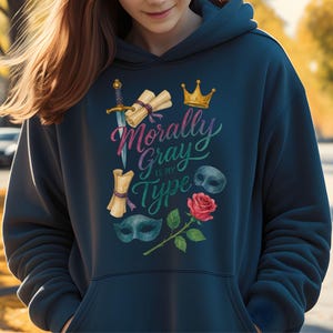 May include: Navy blue hoodie with the phrase "Morally Gray is my Type" in colorful script. The design includes a sword, scrolls, a crown, a rose, and masks. The hoodie has a drawstring hood and a front pocket.