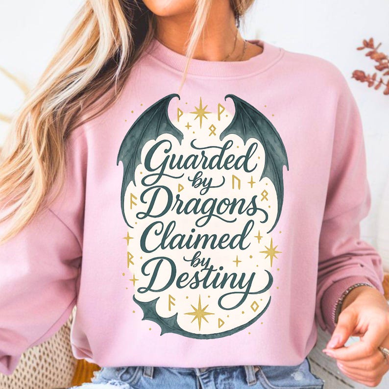 May include: A light pink sweatshirt with the text "Guarded by Dragons, Claimed by Destiny" in a stylized font. The design includes dragon wings and star accents, creating a fantasy theme.