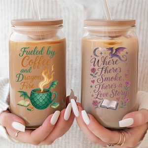 May include: Two glass tumblers with wooden lids and straws, filled with a light brown beverage. One tumbler reads "Fueled by Coffee and Dragon Fire" with a dragon and coffee cup graphic. The other reads "Where There's Smoke, There's a Love Story" with a dragon and rose graphic.