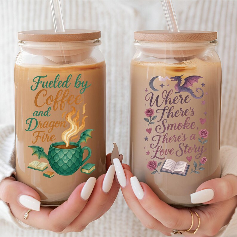 May include: Two glass tumblers with wooden lids and straws, filled with a light brown beverage. One tumbler reads "Fueled by Coffee and Dragon Fire" with a dragon and coffee cup graphic. The other reads "Where There's Smoke, There's a Love Story" with a dragon and rose graphic.