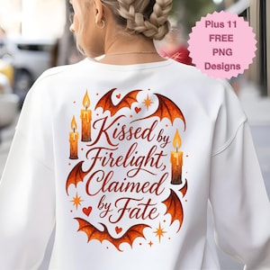 May include: White sweatshirt with a design featuring the text "Kissed by Firelight, Claimed by Fate" in a stylized font. The design includes bat wings, candles, hearts, and stars in shades of orange and brown.