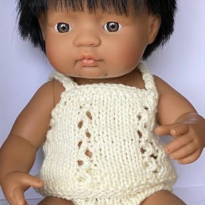 May include: A doll with dark skin and black hair wearing a cream-coloured knitted romper. The romper features a textured pattern with openwork details. The doll has brown eyes and is posed in a seated position.