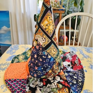 Puede incluir: Un sombrero cónico de tela con un diseño de patchwork que incluye varios patrones como gatos, esqueletos, fantasmas, calabazas, estrellas y escamas coloridas. El sombrero tiene un ala circular ancha y una corona alta y puntiaguda de tela.