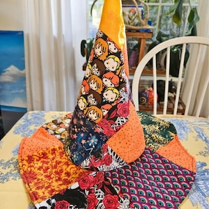 May include: A conical fabric hat featuring a patchwork design of various patterns including cartoon characters, red roses, skulls, and peacock feathers. The top section is solid orange fabric, while the wide brim displays a mix of colorful prints.