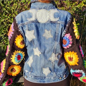 May include: A denim jacket with crochet sleeves. The back of the jacket features a moon and star design. The sleeves are black with colorful crochet flower patterns. The jacket is a light blue wash.