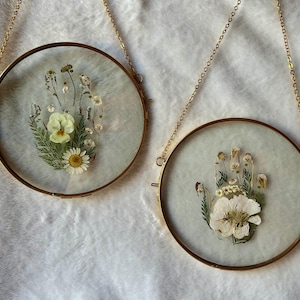 以下が含まれることがあります： 金色のフレームに入った、押し花のアート作品が2つ、金のチェーンで吊るされています。各作品は、デイジーやパンジーなどのドライフラワーを手の形に配置し、半透明の背景に配置しています。