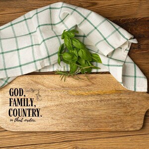 May include: Wooden cutting board with the text "GOD, FAMILY, COUNTRY. in that order." A green and white checkered kitchen towel and fresh basil and rosemary sprigs are on the board. The board has a handle and is on a wooden surface.