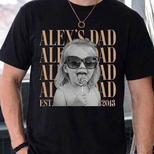 Puede incluir: Camiseta negra con una imagen en sepia de un ni&ntilde;o con gafas de sol y un caramelo. El texto "ALEX'S DAD" est&aacute; impreso verticalmente, con "EST. 2013" debajo.