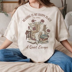 May include: Cream-colored t-shirt with a graphic design featuring a cozy reading nook. The design includes a chair, books, flowers, and the text "Reading is my happy place. Est. 1990s. A Quiet Escape."