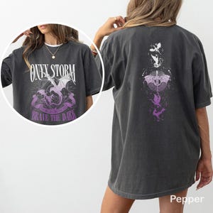 Puede incluir: Camiseta gris oscuro con un dise&ntilde;o de drag&oacute;n, el texto "ONYX STORM" y "BRAVE THE DARK". La parte trasera de la camiseta tiene un dise&ntilde;o a juego con dragones en blanco y morado. La camiseta es de corte holgado.
