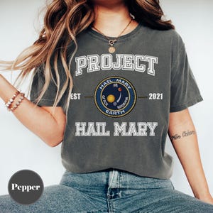 May include: Dark grey t-shirt with the text "PROJECT HAIL MARY" in white block letters. A circular emblem features the words "HAIL MARY", "GRACE EARTH", and "WHOAHIA" around a yellow and red planet design. The year "2021" is also printed.