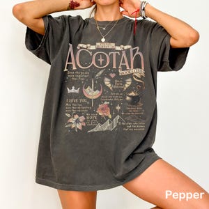May include: Dark gray t-shirt with the text "ACOTAR BOOKS CLUB" and various graphics, including a crown, sword, and floral elements. The shirt also features quotes and the word "Pepper" at the bottom.