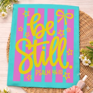 May include: A teal and pink decorative towel with the words "be still" in yellow script. The towel has vertical pink stripes and yellow stars and a bow. The text "Psalm 46:10" is below the main text.