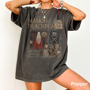 May include: Dark gray t-shirt with the text "MANON BLACKBEAK" in a distressed font. The shirt features a graphic design with a woman in a red cloak, a dragon, and the words "Wind Cleaver" and "Until The Darkness Claims Us."