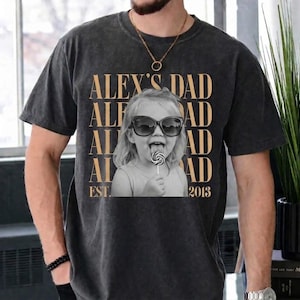 Puede incluir: Camiseta gris oscuro con una foto en blanco y negro de un ni&ntilde;o con gafas de sol y un caramelo. El texto dorado "ALEX'S DAD" est&aacute; impreso encima de la foto, con "EST. 2013" debajo.