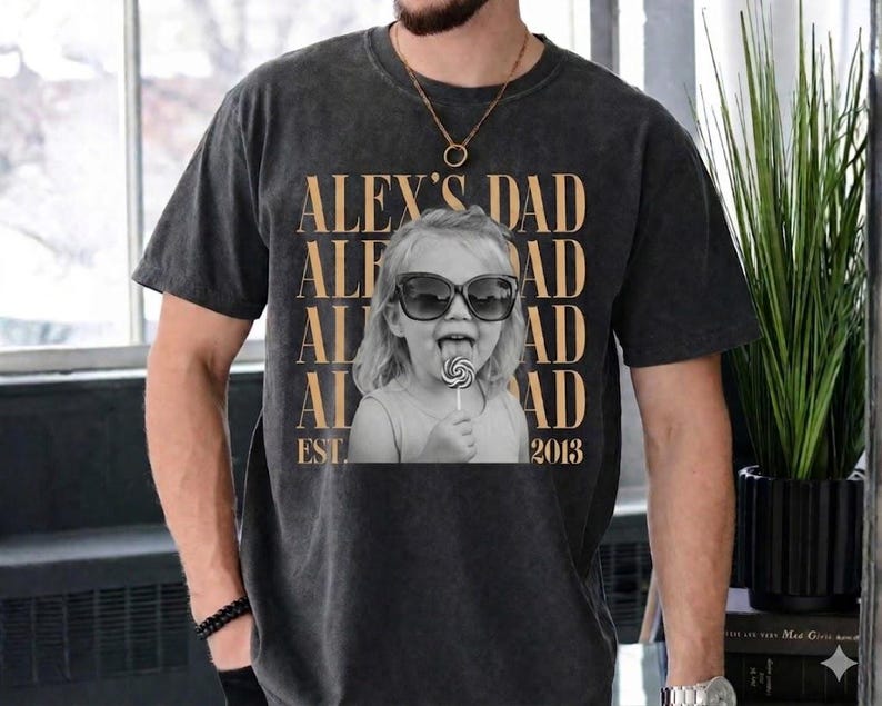 Puede incluir: Camiseta gris oscuro con una foto en blanco y negro de un ni&ntilde;o con gafas de sol y un caramelo. El texto dorado "ALEX'S DAD" est&aacute; impreso encima de la foto, con "EST. 2013" debajo.