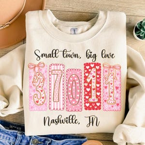 May include: A cream-coloured sweatshirt featuring the phrase "Small town, big love" above the numbers "37011" in pink and red. The words "Nashville, TN" are printed below. The design includes hearts and bows.