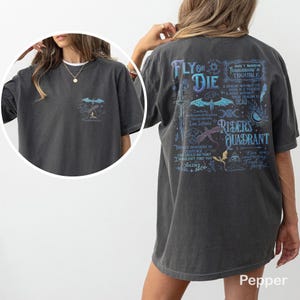 May include: Dark gray t-shirt with a graphic on the front pocket and back. The back graphic features a dragon, sword, and text that reads "FLY OR DIE" and "RIDERS QUADRANT". The shirt is a relaxed fit.
