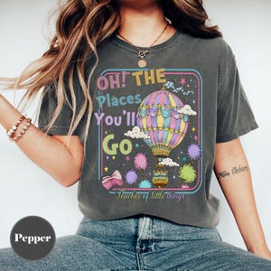 May include: Dark grey t-shirt with a colorful graphic featuring a hot air balloon, clouds, and the text "Oh! The Places You'll Go." The design includes stars and the phrase "Teacher of little things."