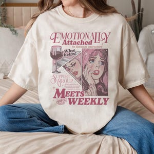 Puede incluir: Camiseta color crema con un diseño gráfico. El diseño presenta el texto "Emotionally Attached to fictional characters" y una ilustración de una mujer llorando y una copa de vino. También se incluyen las palabras "Support Group Meets Weekly".