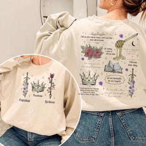Puede incluir: Sudadera beige con varios gráficos y texto. La parte delantera presenta una espada, una corona y una flecha con las palabras "Powerless", "Fearless" y "Reckless". La parte trasera tiene rosas, un cucharón de miel y el texto "Call us ever call me crazy I don't care just..."