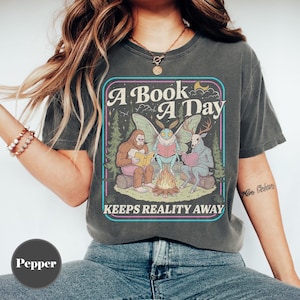 May include: Dark grey t-shirt with a graphic featuring a Bigfoot, a moth, and a deer reading books around a campfire. The text reads "A Book A Day Keeps Reality Away" in a colorful, outlined font.