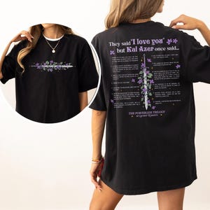 Puede incluir: Camiseta negra con un dise&ntilde;o floral y de espada en la parte delantera. La parte trasera de la camiseta presenta el texto "They said 'I love you' but Kai Azer once said..." y una lista de frases. La camiseta es parte de "The Powerless Trilogy" de Lauren Roberts.