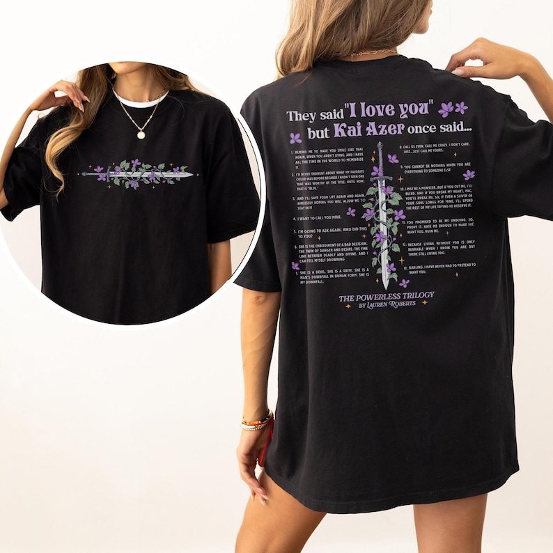 Puede incluir: Camiseta negra con un dise&ntilde;o floral y de espada en la parte delantera. La parte trasera de la camiseta presenta el texto "They said 'I love you' but Kai Azer once said..." y una lista de frases. La camiseta es parte de "The Powerless Trilogy" de Lauren Roberts.
