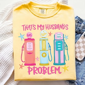 Könnte beinhalten: Gelbes Comfort Colors T-Shirt mit dem pinkfarbenen Schriftzug "THAT'S MY HUSBANDS PROBLEM". Das Shirt zeigt drei farbenfrohe Tankstellengrafiken. Ein Telefon mit kariertem Gehäuse und Jeans sind im Hintergrund.