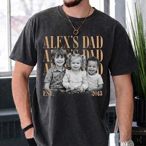 Puede incluir: Camiseta gris oscuro con el texto "ALEX'S DAD" repetido tres veces en color dorado. Una foto en blanco y negro de tres ni&ntilde;os est&aacute; centrada debajo, con "EST. 2013" impreso debajo.