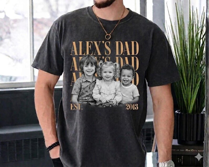 Puede incluir: Camiseta gris oscuro con el texto "ALEX'S DAD" repetido tres veces en color dorado. Una foto en blanco y negro de tres ni&ntilde;os est&aacute; centrada debajo, con "EST. 2013" impreso debajo.