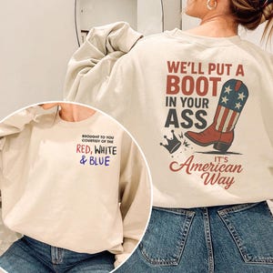 May include: Cream-colored sweatshirt with the text "WE'LL PUT A BOOT IN YOUR ASS" and "It's American Way" in red, white, and blue. The design includes a cowboy boot with the American flag. The front of the sweatshirt says "BROUGHT TO YOU COURTESY OF THE RED, WHITE & BLUE."