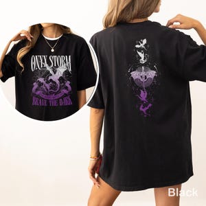 Puede incluir: Camiseta negra con un dise&ntilde;o gr&aacute;fico frontal que presenta un drag&oacute;n, las palabras "ONYX STORM" y "BRAVE THE DARK" en blanco y morado. La parte trasera de la camiseta tiene un dise&ntilde;o de drag&oacute;n a juego en morado y blanco.