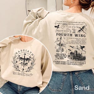 May include: Sand-coloured sweatshirt with graphics from "Fourth Wing." The front has a circular design with a dragon and "Basgiath War College." The back features quotes and illustrations, including dragons and text. The sweatshirt is made of a soft material, perfect for fans of the book.