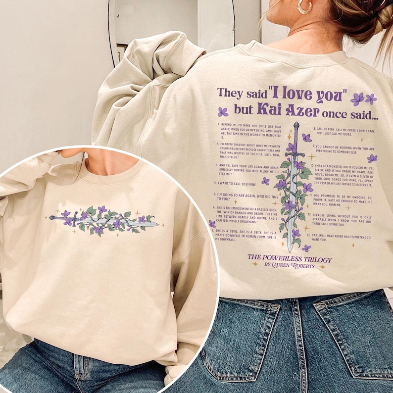 Puede incluir: Una sudadera beige con el texto "They said 'I love you' but Kai Azer once said..." y un dise&ntilde;o de espada con flores moradas. La sudadera tambi&eacute;n presenta texto de "The Powerless Trilogy" de Lauren Roberts.